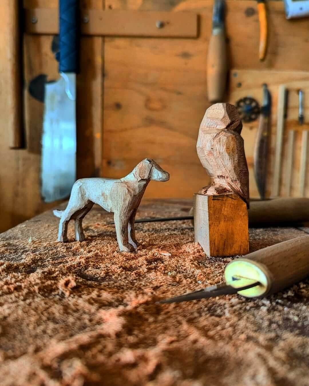 A dog and an owl carved from wood with carving tools in the background and laying on the table, surrounded by wood shavings